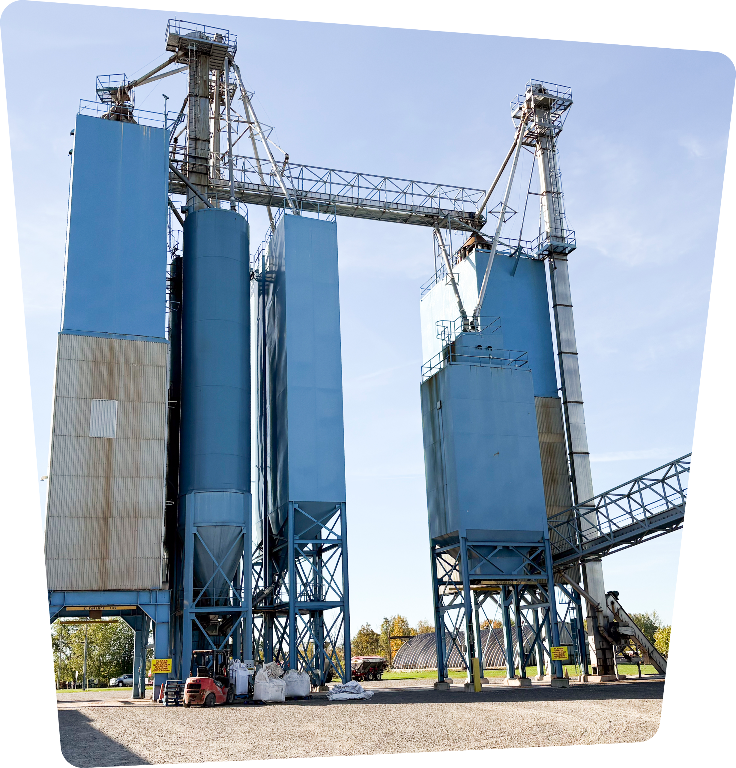 Tall blue feed mill silos and conveyors at an agricultural processing site.